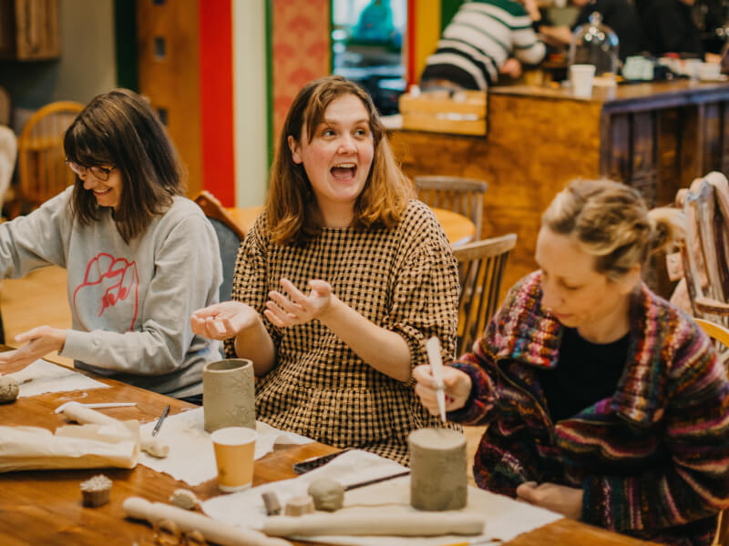 Several smiling people sit at a table making clay pots with engravings on them